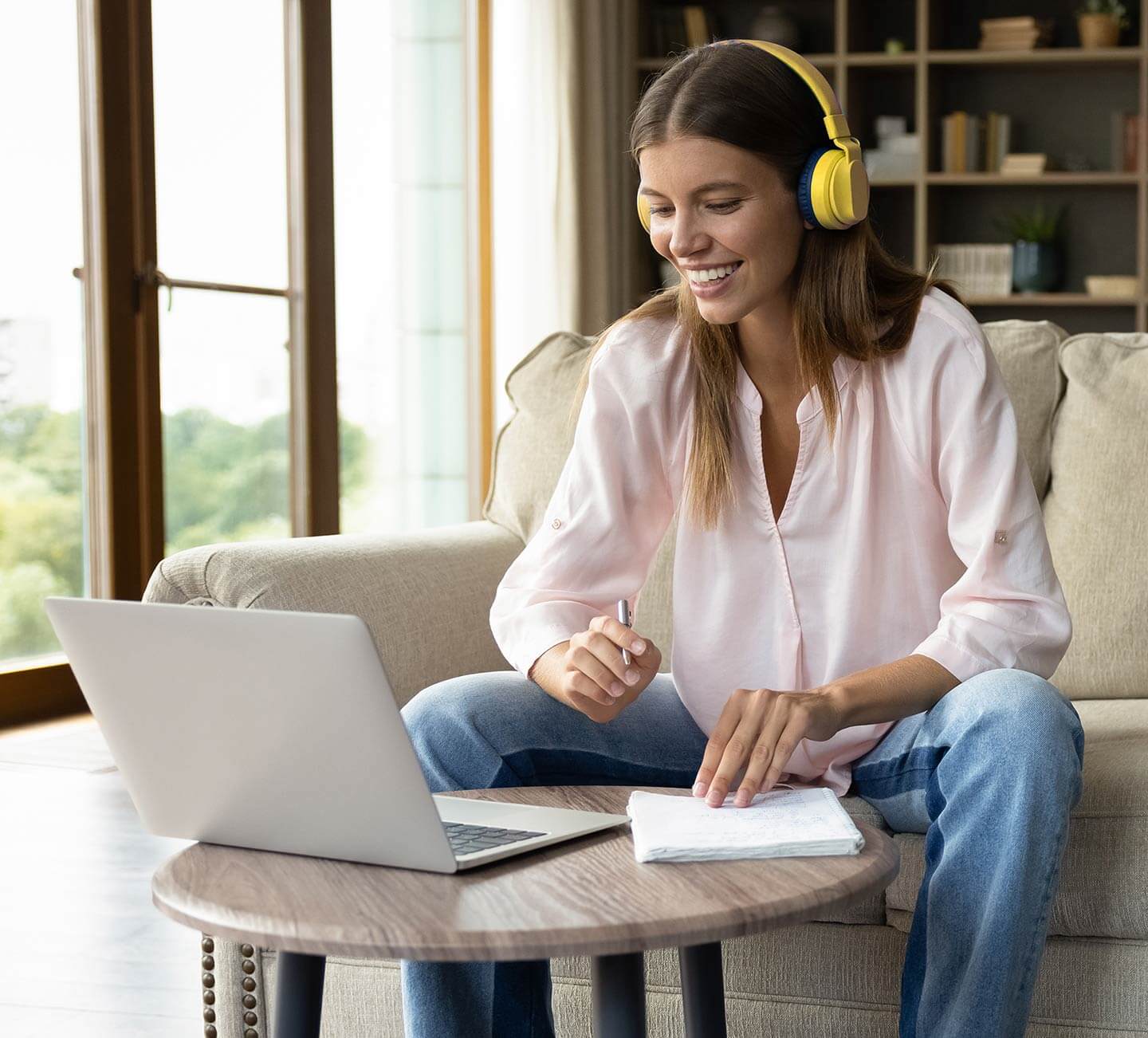 Smiling woman wearing headphones as she takes notes while viewing a laptop